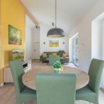 Essbereich mit runden Tisch und grünen Stühlen. Im Hintergrund Wohnbereich mit Sofa und Fenster.