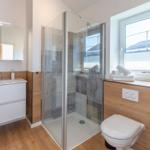 Badezimmer mit Dusche, Waschbecken und Toilette. Holzoptik und Fenster mit Blick.