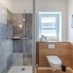 Badezimmer mit Dusche, Toilette und Fenster. Wandfliesen mit Muster. Holzverkleidung an Wänden.