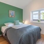 Schlafzimmer mit Bett, grüner Wand, Nachttisch und Fenster mit Pflanze.