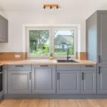 Küche mit grauen Schränken, Holzoberflächen und Fensterblick auf eine Scheune.