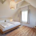 Schlafzimmer mit Doppelbett, Holzboden und Dachschrägen. Fenster mit Vorhängen und Sitzgelegenheit.