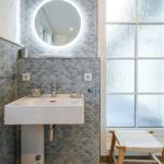 Bad mit Waschbecken, runden Spiegel und gefliesten Wänden. Fenster mit Sichtschutz und Holzstuhl.