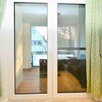 Durchgehende Glastüren führen auf eine Terrasse mit Sitzgelegenheiten. Grüne Vorhänge rahmen den Eingang ein.