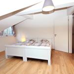 Schlafzimmer mit Dachschrägen, weißem Bett und Holzboden. Fenster mit Blick nach draußen.