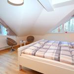 Schlafzimmer mit Dachschrägen, Bett, zwei Sessel und Fenstern mit Blick auf Wald.