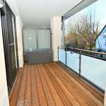 Terrasse mit Holzdielen, Glasgeländer und Außenspeicher. Blick auf Bäume und Nachbarhäuser.