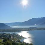 Panoramablick auf einen See mit Bergen im Hintergrund und Sonne am Himmel.