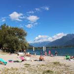 Strand mit Steinen, Menschen, Bäumen und Bergen im Hintergrund. Blauer Himmel mit weißen Wolken.