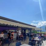Terrasse mit Tischen und Stühlen, Blick auf See und Berge unter blauem Himmel.
