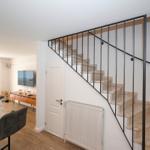 Treppe mit Metallgeländer führt zur oberen Etage. Im Hintergrund Wohnbereich mit Fernseher und Holzboden.