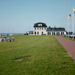 Grüner Strandbereich mit Strandkörben, Weg und Gebäude im Hintergrund.