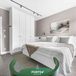 Schlafzimmer mit weißem Bett, Kleiderschrank und Wandbild. Durchgang zu Wohnbereich mit Balkon.