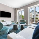 Wohnzimmer mit Sofa, Sesseln, Couchtisch und TV. Durchgang zur Terrasse mit Blick auf Gebäude.