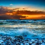 Wellen brechen auf einem steinigen Strand bei Sonnenuntergang mit dramatischem Himmel.