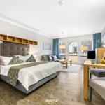 Schlafzimmer mit Bett, Wohnbereich und Essplatz. Holzboden und große Fenster.