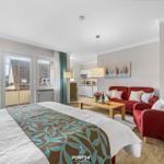 Ein Schlafzimmer mit Blick auf Balkon, Wohnbereich mit rotem Sofa und Küchenbereich.