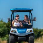 Zwei Personen fahren in einem blauen Golfcart auf einer Straße.