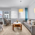 Wohnzimmer mit Sofa, Sessel und Tisch. Fenster mit Vorhängen und Deckenleuchten.