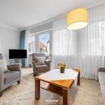 Wohnzimmer mit Sofa, Sesseln, Tisch und TV. Große Fenster mit weißen Vorhängen.