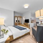 Schlafzimmer mit Bett, Nachttisch und Lampe. Küche und Wohnbereich im Hintergrund.