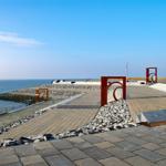 Steinpflasterter Strandweg mit roten Metallskulpturen und Blick auf das Meer.