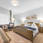 Schlafzimmer mit Holzbett, Nachttischen und Sitzgelegenheit. Fenster mit weißen Vorhängen.