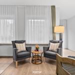 Zwei graue Sessel stehen vor einem Fenster mit weißen Vorhängen. Zwischen ihnen steht ein runder Tisch.