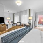 Schlafzimmer mit Bett, TV, Sitzgelegenheit und Balkon. Holzmöbel und Fenster mit Vorhängen.