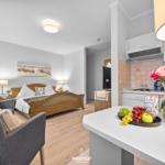 Wohnbereich mit Schlafzimmer und Küche. Auf der Kücheninsel stehen Obst und Blumen.