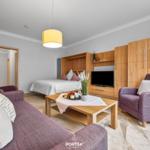 Wohnbereich mit Sofa, Sessel und Bett. Holzmöbel und Fernseher sind sichtbar.