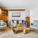 Wohnzimmer mit Sofa, zwei Sesseln und Glas-Tisch. Holzmöbel und Wandbild mit Segelbooten.