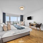 Schlafzimmer mit Balkon, TV und Esstisch. Holzboden und große Fenster.