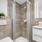 Bad mit Dusche, Waschbecken und Toilette. Fliesenwand und Spiegel mit Beleuchtung.