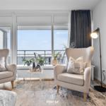 Zwei Sessel stehen vor Fenstern mit Meerblick. Tisch, Teppich und Stehlampe sind vorhanden.