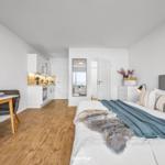 Schlafzimmer mit Bett, Nachttisch und Wandspiegel. Küchenbereich und Esstisch sind sichtbar.