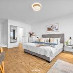 Schlafzimmer mit Bett, Nachttisch und Spiegel. Küchenbereich und Esstisch sind sichtbar.