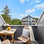 Balcony with wooden table, chairs, and fruit bowl. View of a house and trees under blue sky.