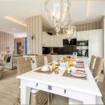 Esszimmer mit weißem Tisch und Rattanstühlen, Küche im Hintergrund, Wohnzimmer mit Sofa und Wandtapete.