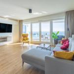 Wohnzimmer mit Sofa, Fernseher und Meerblick durch große Fenster.