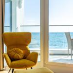 Gelber Sessel mit Kissen vor Fenster mit Meerblick und Balkonmöbeln.