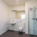 Badezimmer mit Dusche, Waschbecken und Toilette. Weiße Fliesen und graue Bodenfliesen.