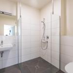 Badezimmer mit Dusche, Waschbecken und Toilette. Weiße Fliesen und graue Bodenfliesen.