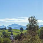 Weitläufiger Blick auf grüne Hügel mit Kirche und Bergen im Hintergrund unter blauem Himmel.