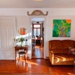 Ein Wohnzimmer mit braunem Sofa, Holzboden und Kronleuchter. Durchgang zu Küche mit Fenster.