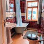 Bad mit freistehender Badewanne, Waschtisch und Toilette. Rote Fliesen und Holzfenster.