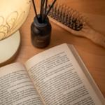 Ein offenes Buch liegt auf einem Holztisch neben einer Duftflasche und einem Haarbürsten.