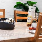 Wooden table with chairs, bread, drinks, and plant in background.