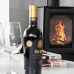 A table with a wine bottle and glasses in front of a burning wood stove.