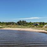 Strandufer mit Häusern und Windkraftanlagen im Hintergrund unter blauem Himmel.